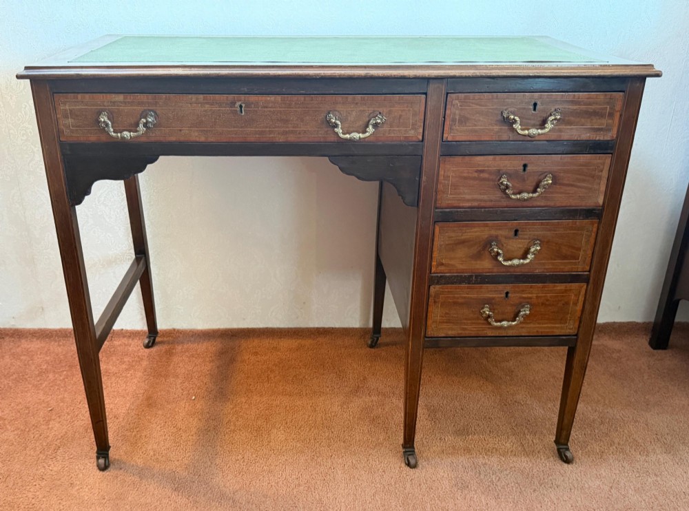 An antique mahogany kneehole writing desk featuring a green leather top surface, inlaid stringing, five drawers with brass handles, and square tapered legs on castors. View II