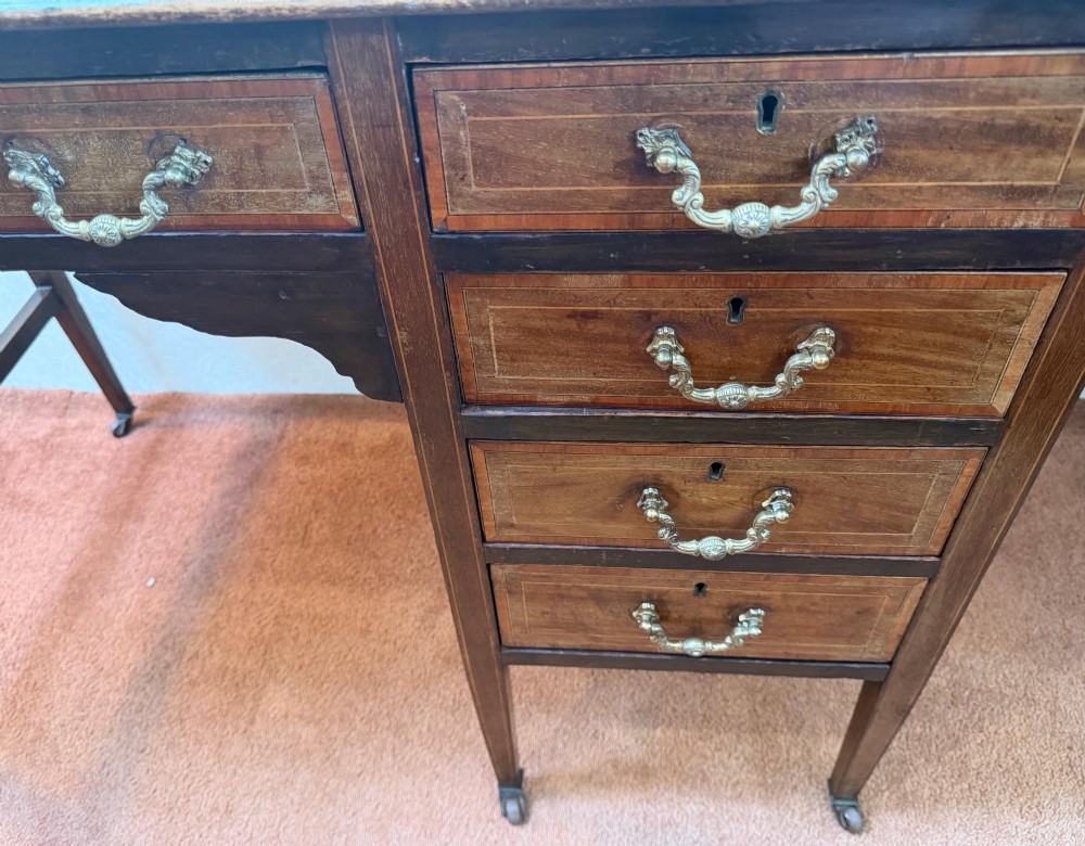 An antique mahogany kneehole writing desk featuring a green leather top surface, inlaid stringing, five drawers with brass handles, and square tapered legs on castors. View III