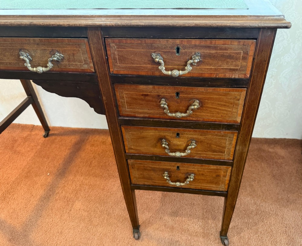 An antique mahogany kneehole writing desk featuring a green leather top surface, inlaid stringing, five drawers with brass handles, and square tapered legs on castors. View IV
