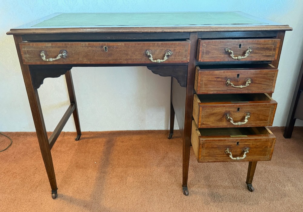 An antique mahogany kneehole writing desk featuring a green leather top surface, inlaid stringing, five drawers with brass handles, and square tapered legs on castors. View V