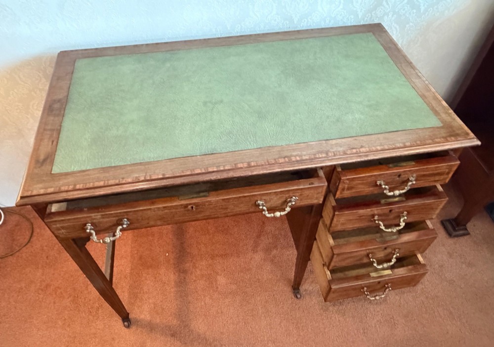 An antique mahogany kneehole writing desk featuring a green leather top surface, inlaid stringing, five drawers with brass handles, and square tapered legs on castors. View VI