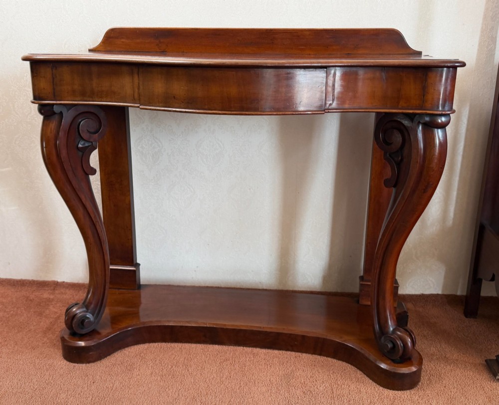 A Victorian mahogany serpentine console table featuring carved S-scroll legs, a low splashback upstand, and a shaped plinth base.