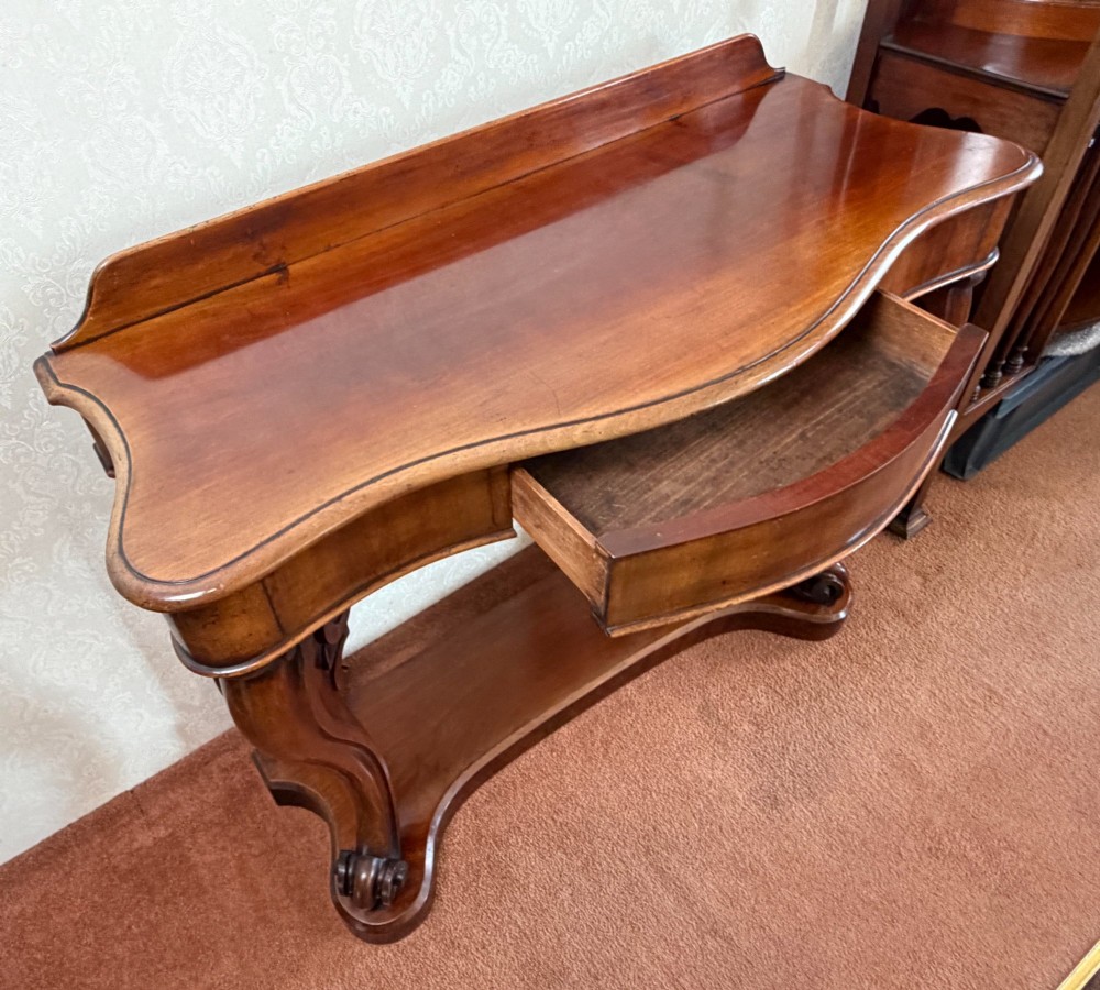 A Victorian mahogany serpentine console table featuring carved S-scroll legs, a low splashback upstand, and a shaped plinth base. View IV
