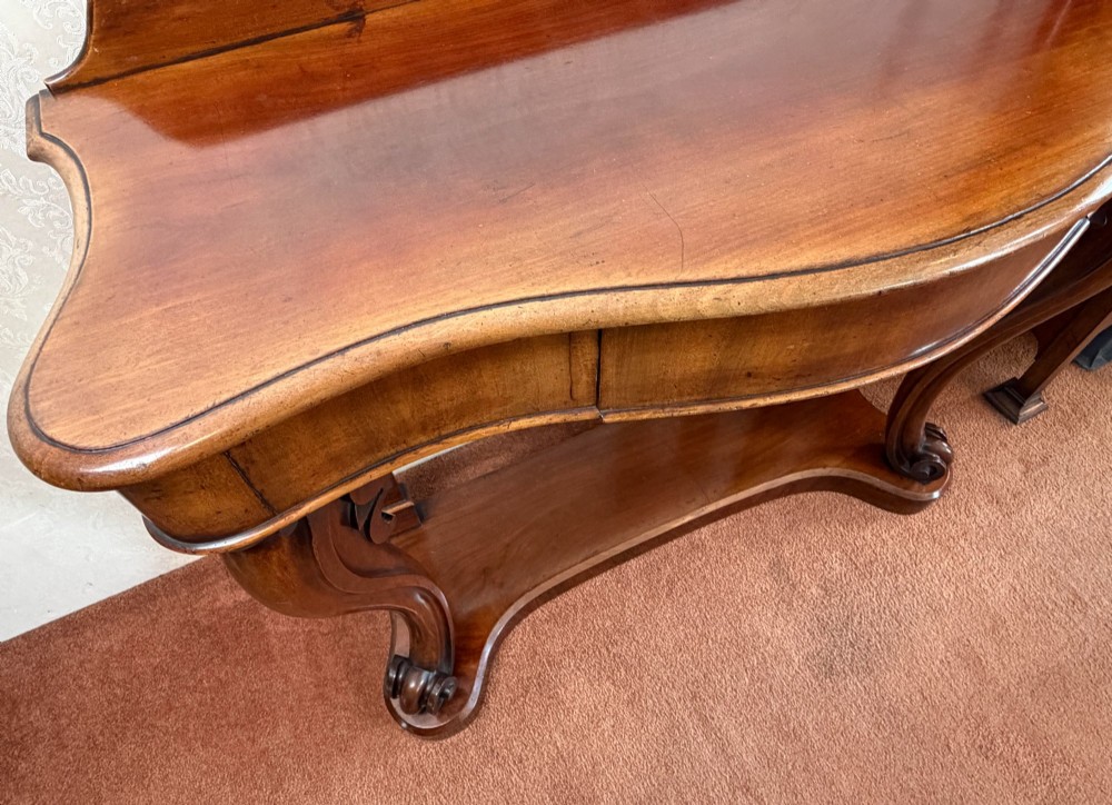 A Victorian mahogany serpentine console table featuring carved S-scroll legs, a low splashback upstand, and a shaped plinth base. View V