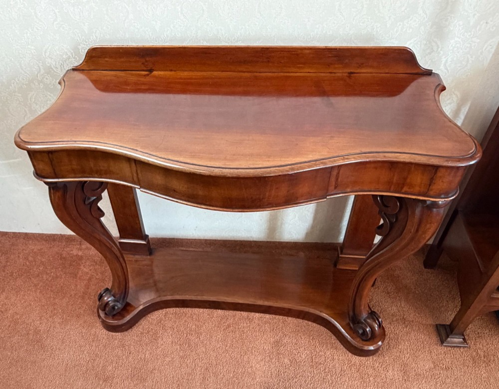 A Victorian mahogany serpentine console table featuring carved S-scroll legs, a low splashback upstand, and a shaped plinth base. View VI