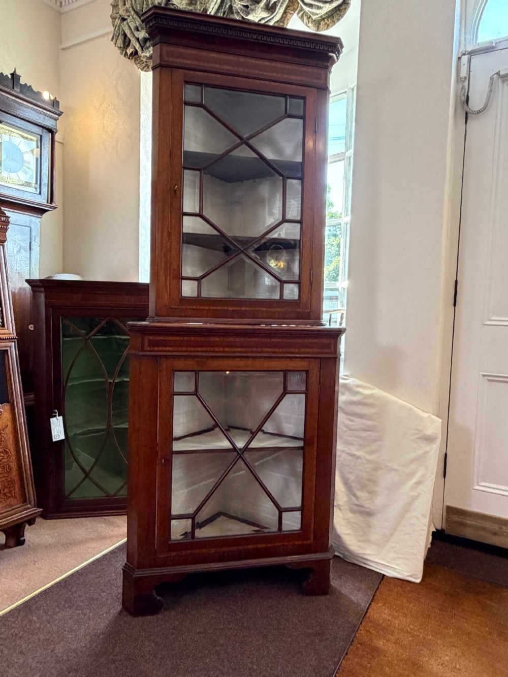 A tall two-tier mahogany corner cabinet featuring geometric glass door panes, satinwood inlay details, and bracket feet.