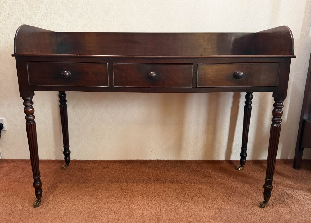 A dark mahogany writing table featuring a raised gallery back, three frieze drawers with turned wooden knobs, turned reeded legs, and brass castors.