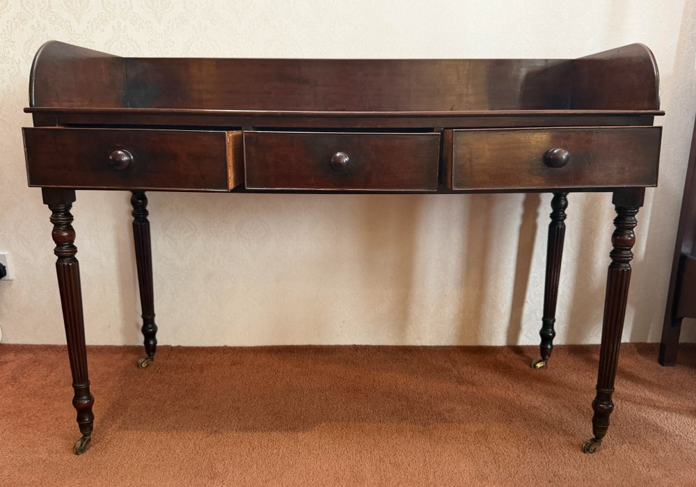 A dark mahogany writing table featuring a raised gallery back, three frieze drawers with turned wooden knobs, turned reeded legs, and brass castors. View II