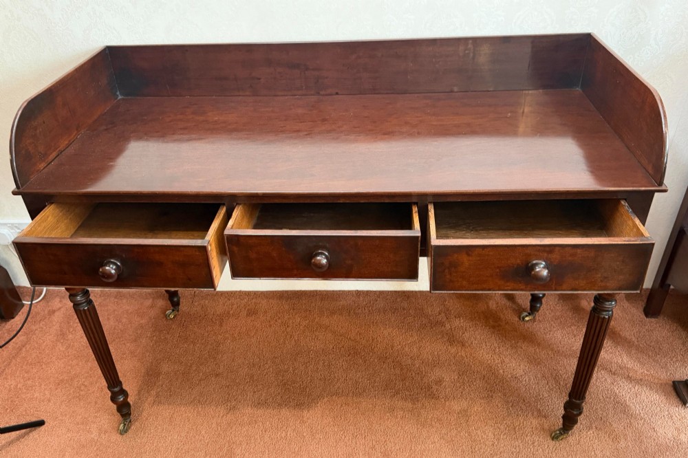 A dark mahogany writing table featuring a raised gallery back, three frieze drawers with turned wooden knobs, turned reeded legs, and brass castors. View III