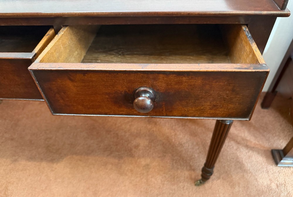 A dark mahogany writing table featuring a raised gallery back, three frieze drawers with turned wooden knobs, turned reeded legs, and brass castors. View IV
