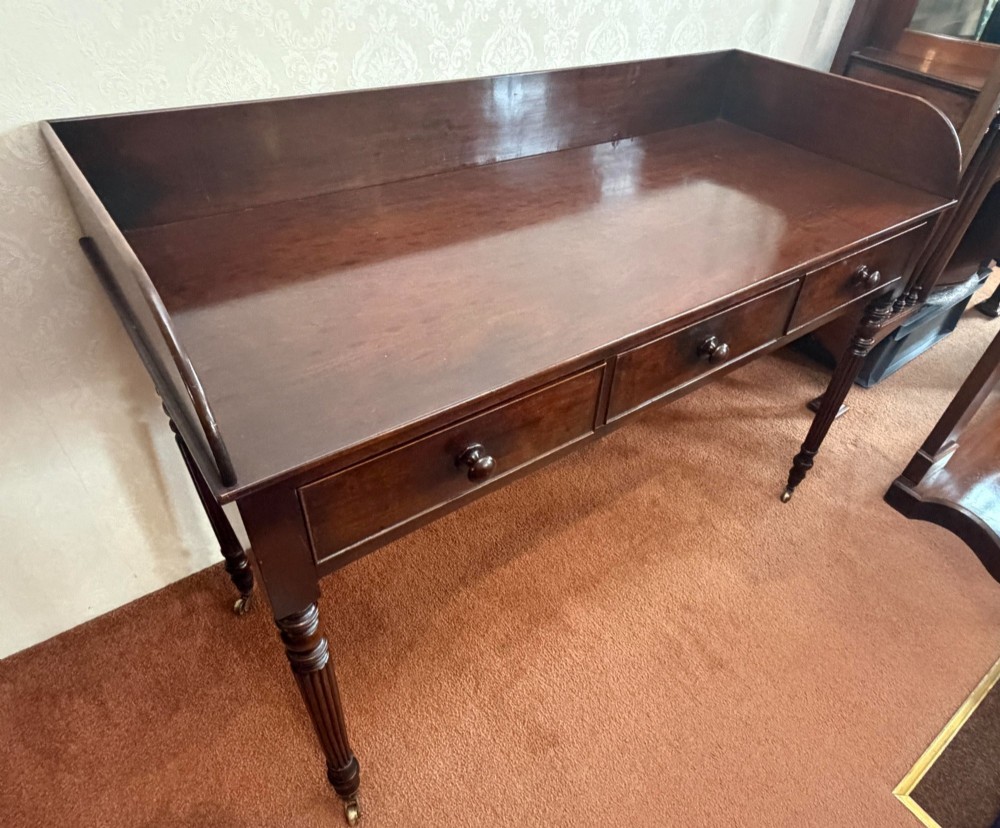 A dark mahogany writing table featuring a raised gallery back, three frieze drawers with turned wooden knobs, turned reeded legs, and brass castors. View VI