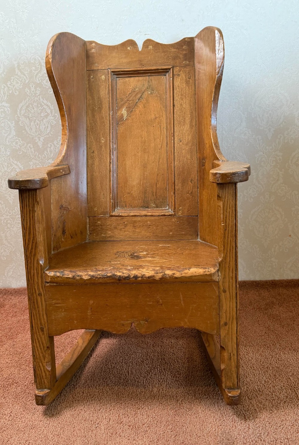 A small wooden antique childs rocking chair featuring solid enclosed panel sides, integrated armrests, and curved wooden rockers. View III