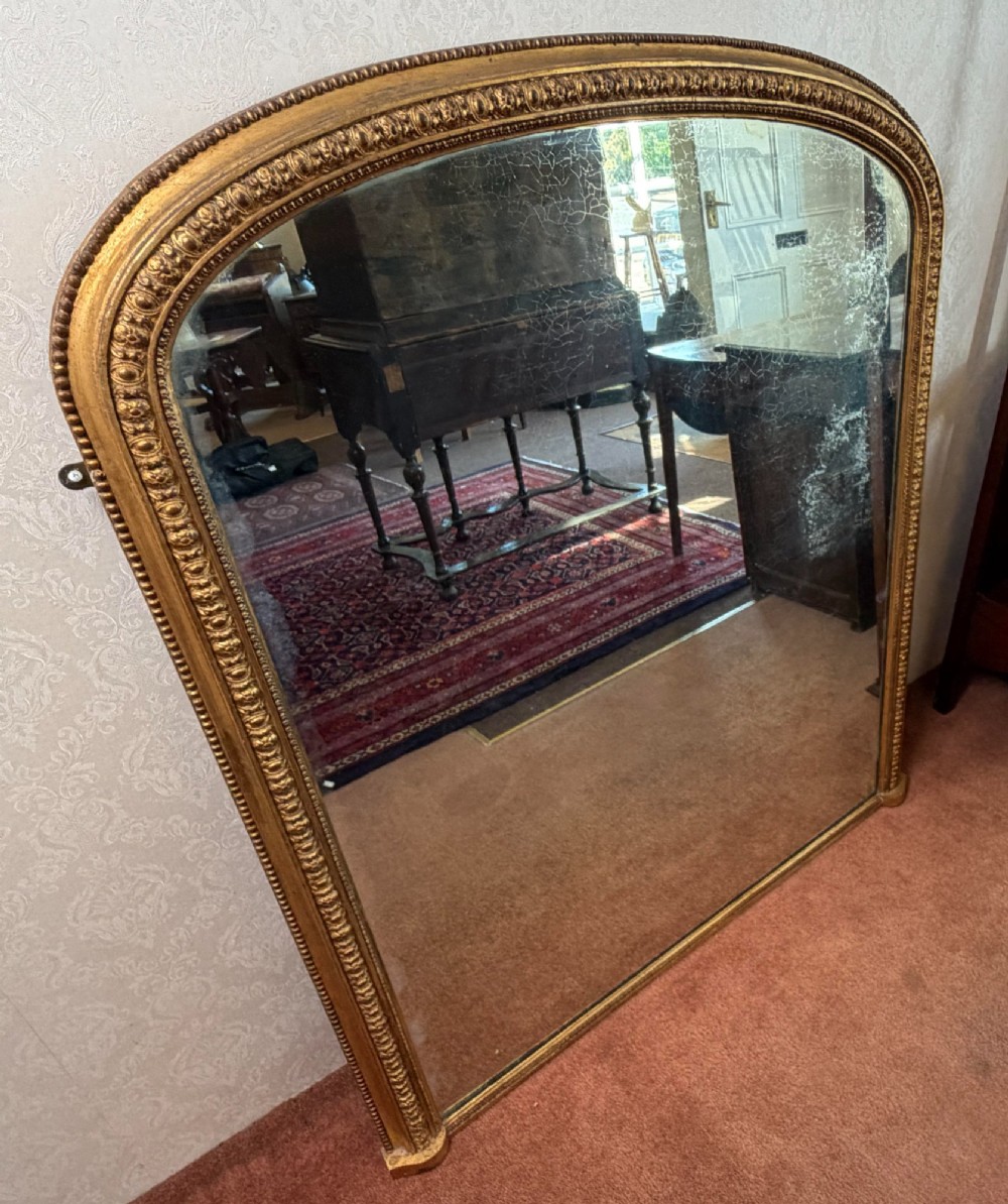 A Victorian arched overmantel mirror featuring a gilded wood frame with beaded moulding and foxed glass plate.