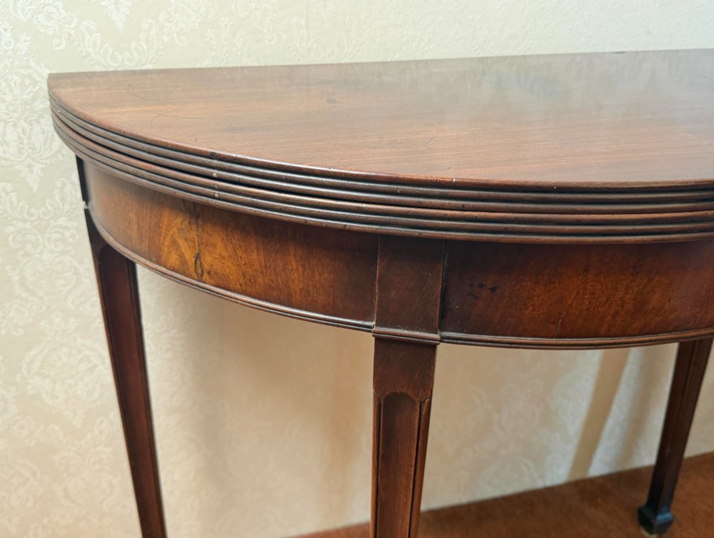 An 18th-century mahogany demi-lune fold-over tea table with tapered reeded legs standing against a wall. View II