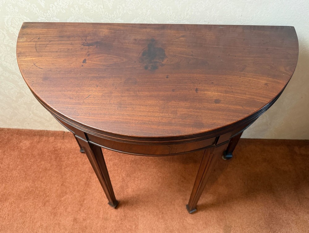 An 18th-century mahogany demi-lune fold-over tea table with tapered reeded legs standing against a wall. View III