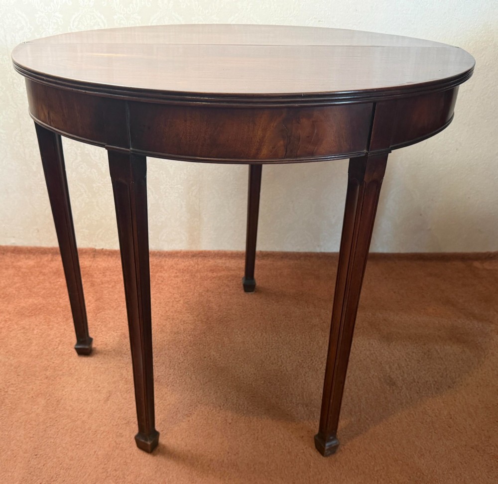 An 18th-century mahogany demi-lune fold-over tea table with tapered reeded legs standing against a wall. View IV