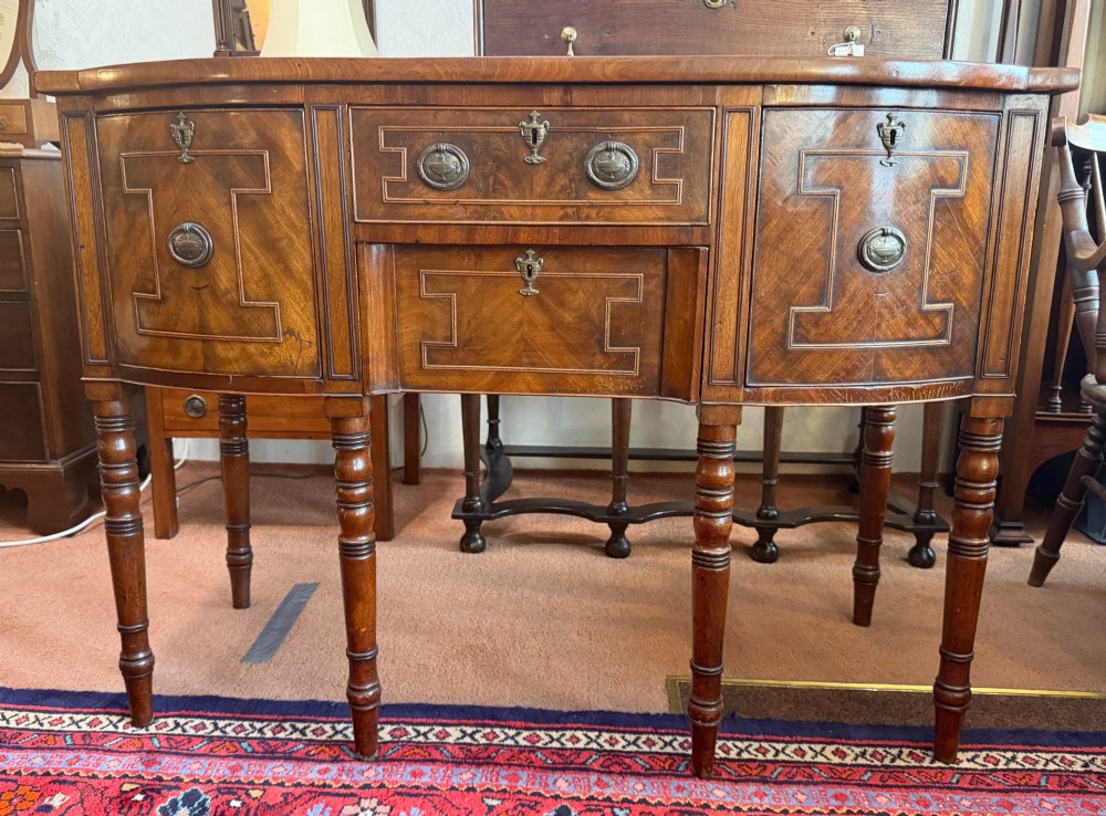 A Regency style mahogany bow-front sideboard featuring geometric string inlay, central drawers flanked by cupboards, brass drop handles, and turned legs.