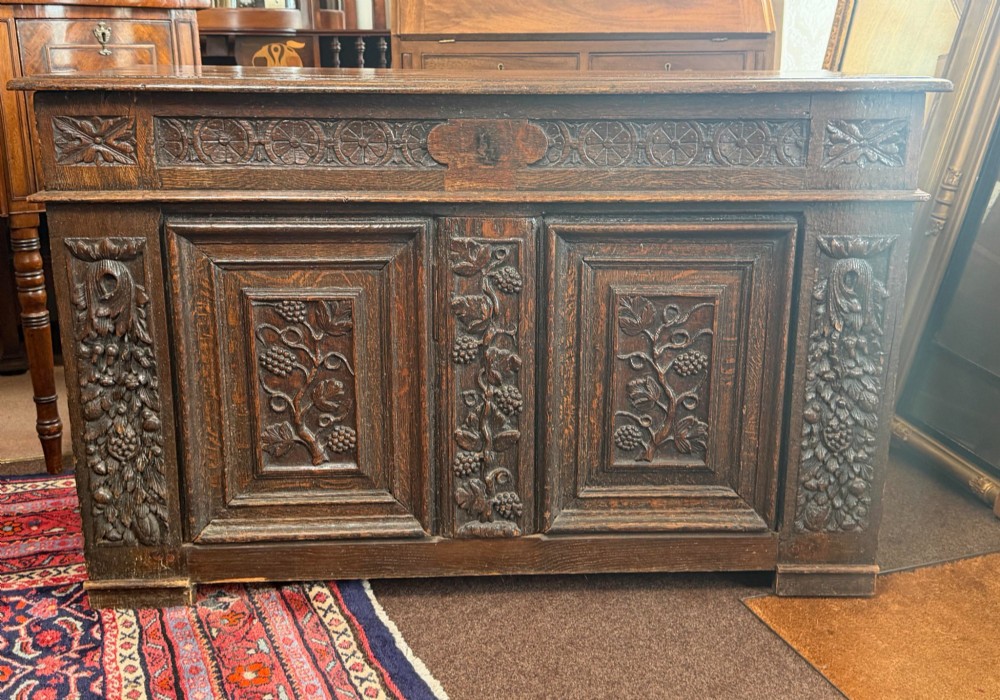 A 17th-century dark oak coffer featuring hand-carved vine and grape motifs across three front panels.