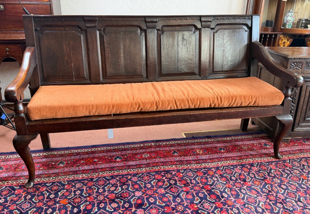 An eighteenth-century dark oak pub settle featuring a four-panel backrest, curved armrests, cabriole legs, and a fitted terracotta seat cushion.