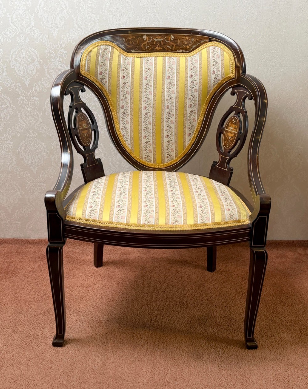 Late 19th century mahogany armchair featuring decorative bone and boxwood urn marquetry, curved arms, and yellow striped floral upholstery.