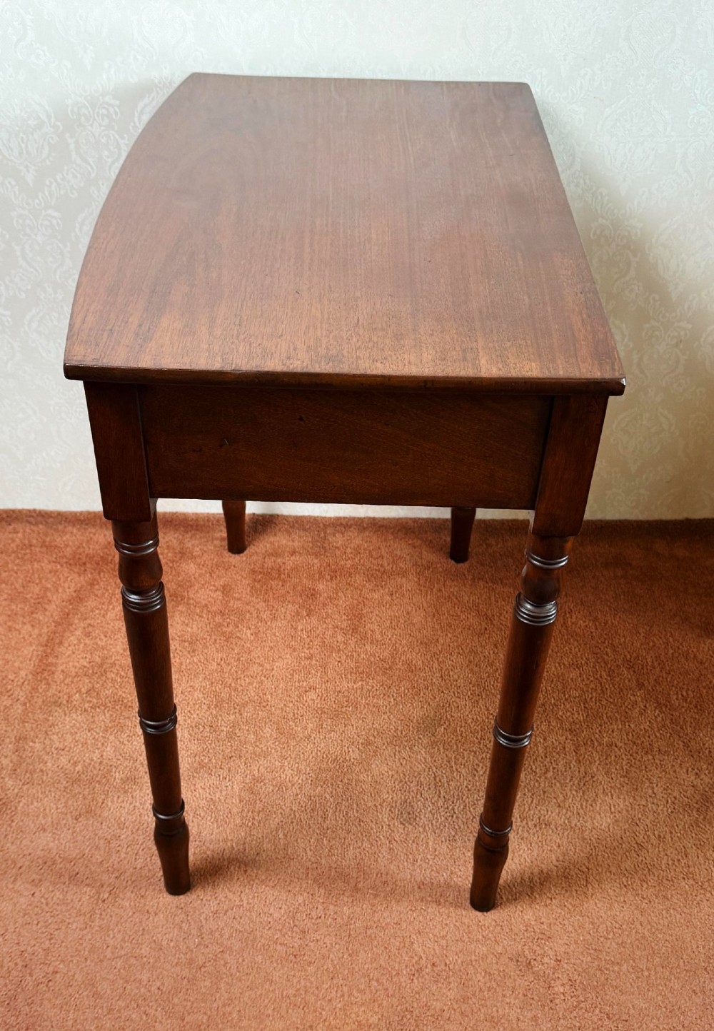 A mahogany bow front side table with two drawers, brass knobs, and slender turned legs. View IV