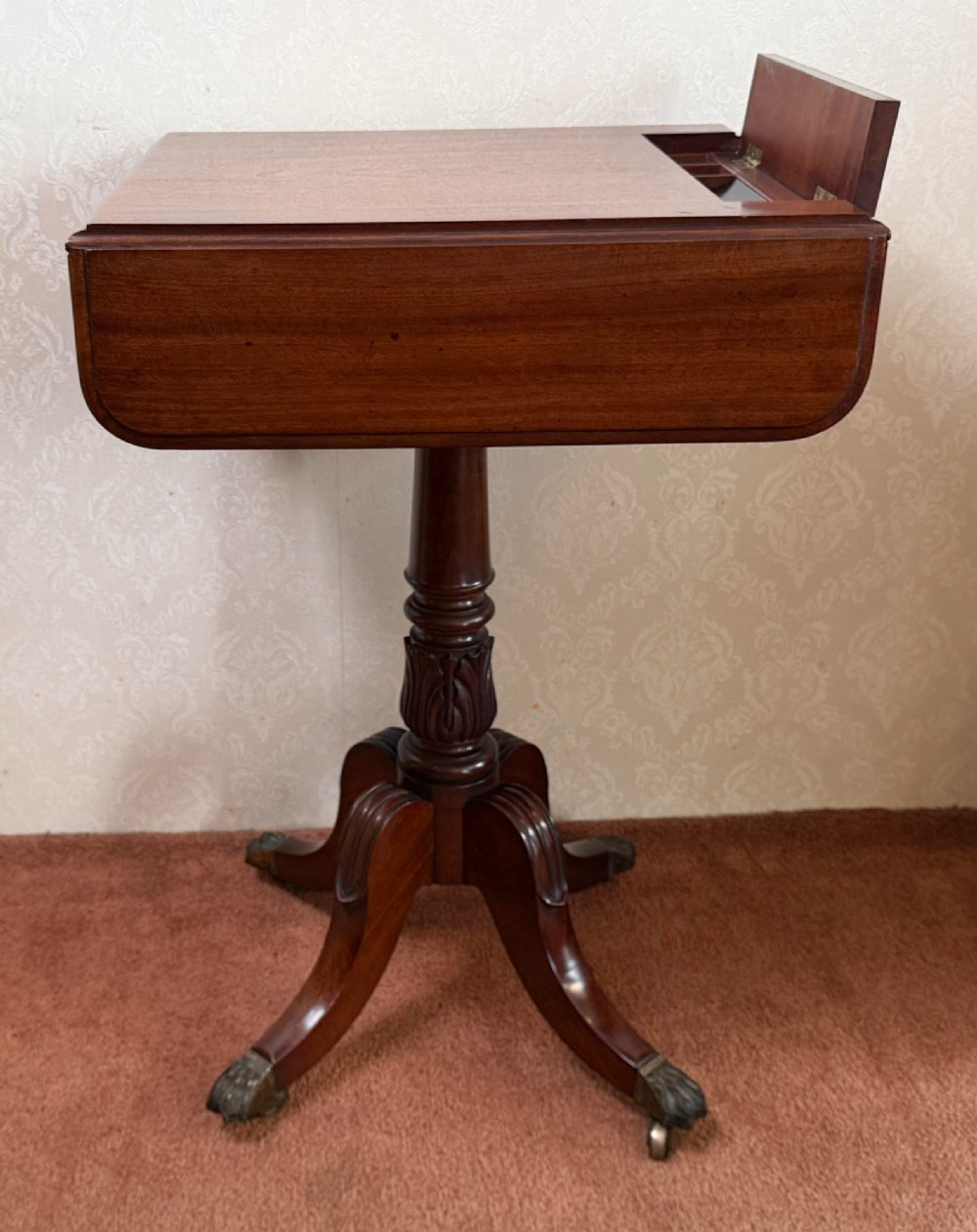 An antique Regency mahogany work table featuring a carved acanthus pedestal column, quadruped splayed legs with brass paw castors, and a hinged lid revealing a fitted compartment.