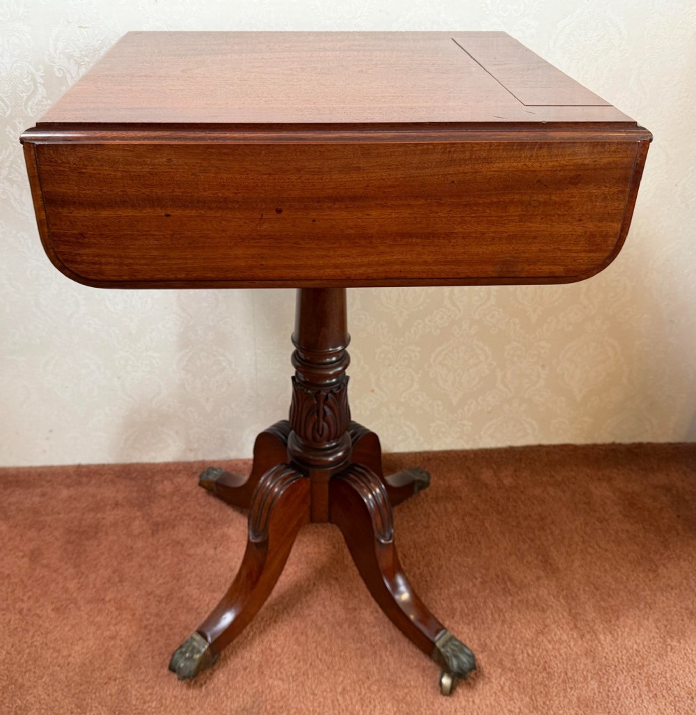 An antique Regency mahogany work table featuring a carved acanthus pedestal column, quadruped splayed legs with brass paw castors, and a hinged lid revealing a fitted compartment. View III