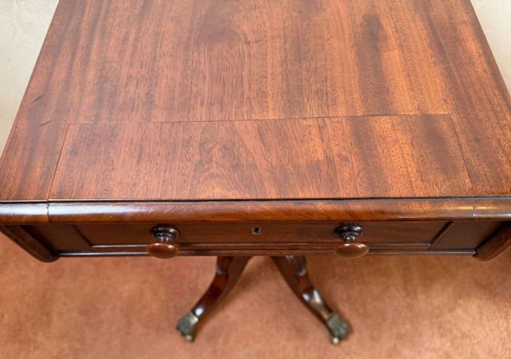 An antique Regency mahogany work table featuring a carved acanthus pedestal column, quadruped splayed legs with brass paw castors, and a hinged lid revealing a fitted compartment. View VI