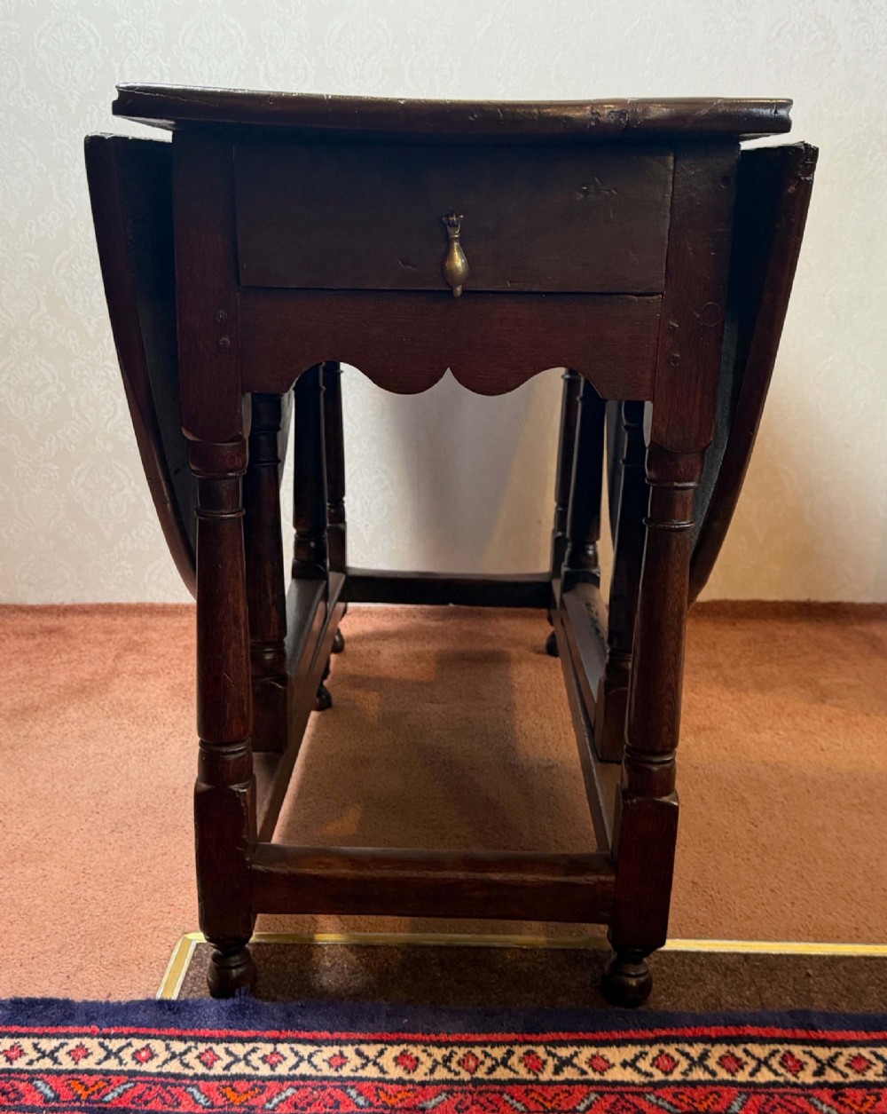 A 17th-century dark oak gateleg table shown with drop leaves lowered, featuring turned legs, a stretcher base, and a drawer with a brass teardrop pull handle.