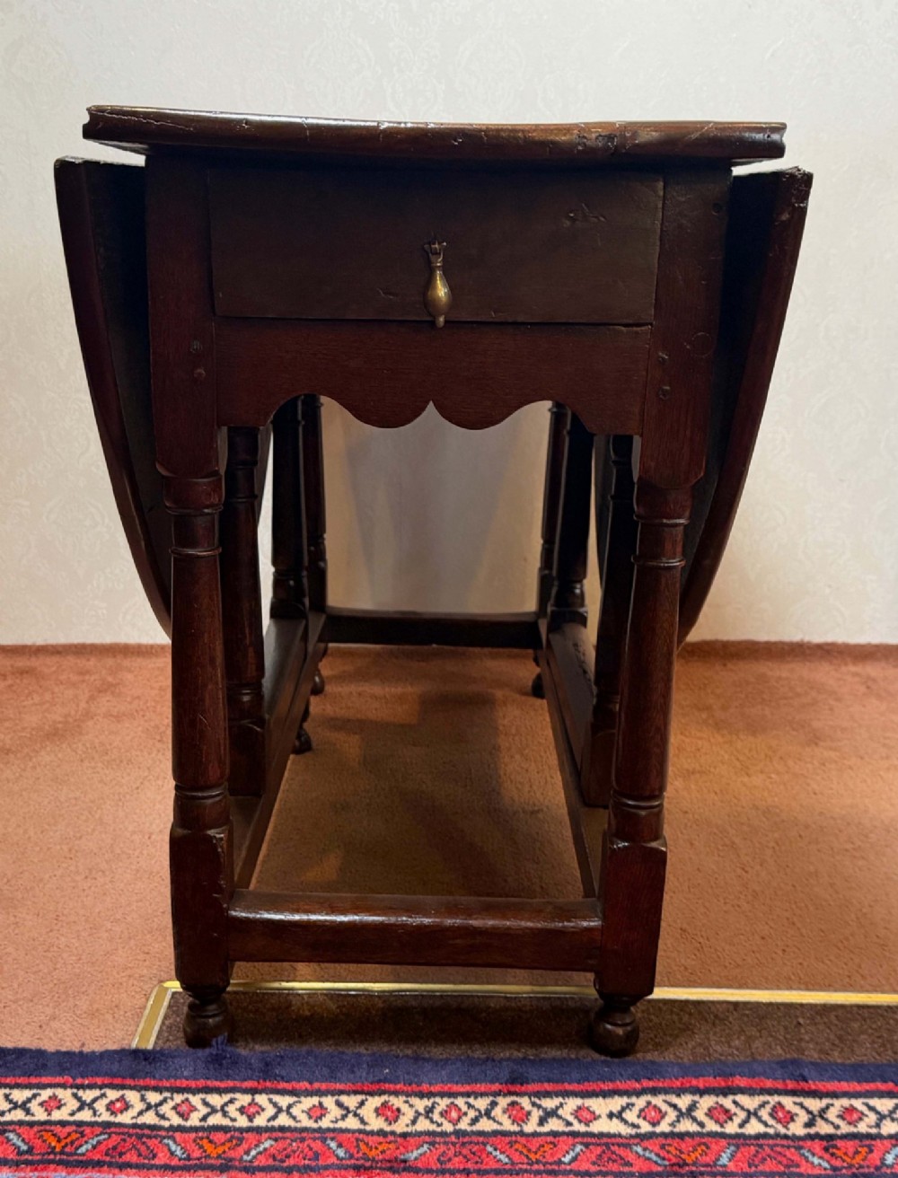 A 17th-century dark oak gateleg table shown with drop leaves lowered, featuring turned legs, a stretcher base, and a drawer with a brass teardrop pull handle. View II