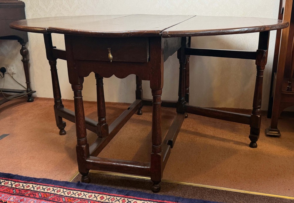 A 17th-century dark oak gateleg table shown with drop leaves lowered, featuring turned legs, a stretcher base, and a drawer with a brass teardrop pull handle. View VII