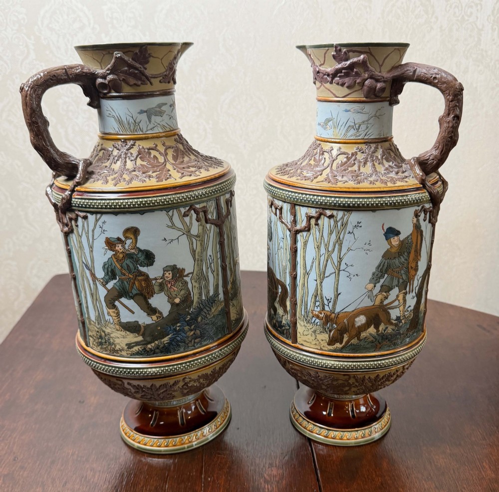 A pair of 19th-century Mettlach stoneware vases featuring sculpted branch handles, oak leaf relief decoration, and incised pictorial hunting scenes in blue and brown.