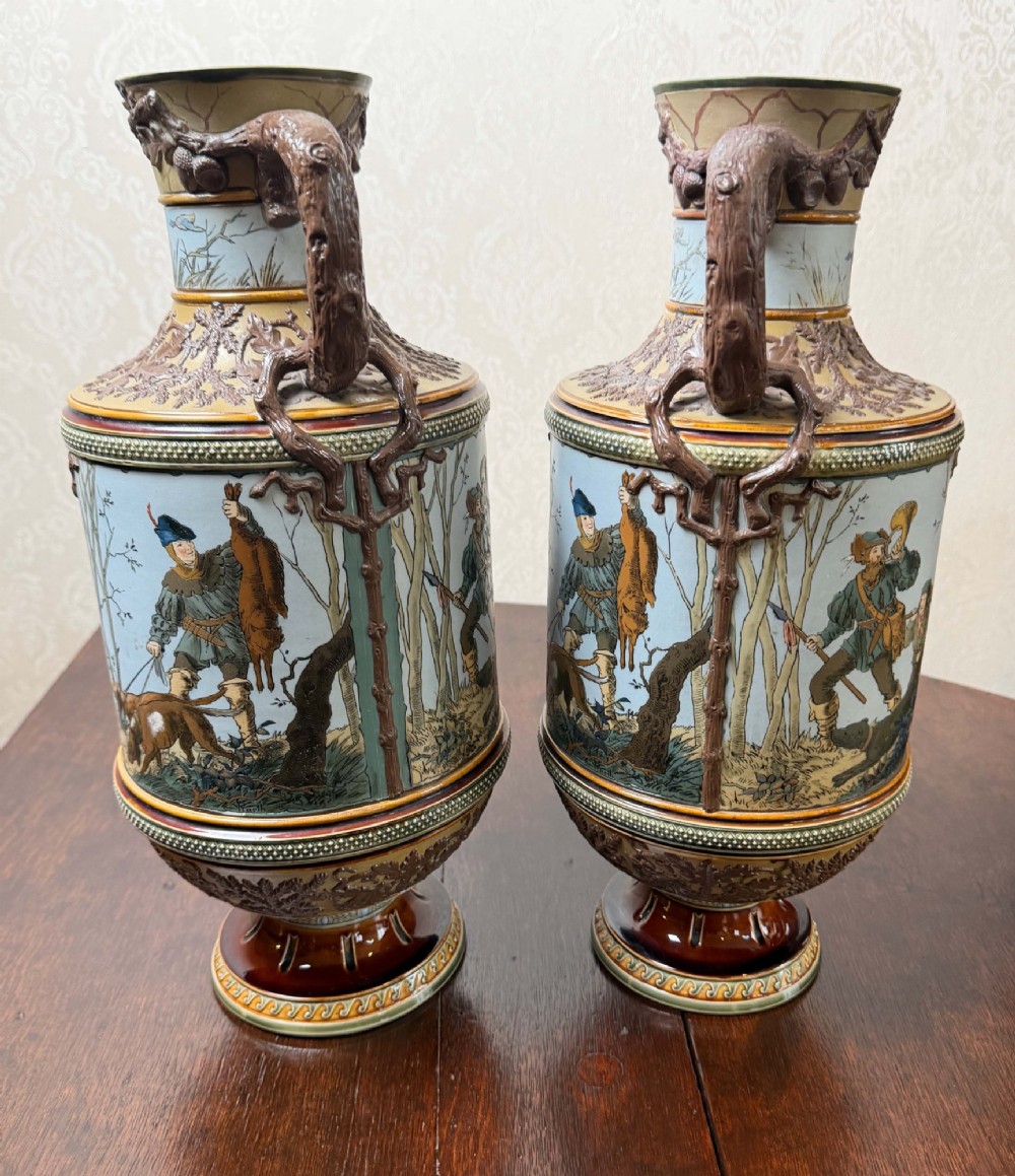 A pair of 19th-century Mettlach stoneware vases featuring sculpted branch handles, oak leaf relief decoration, and incised pictorial hunting scenes in blue and brown. View II