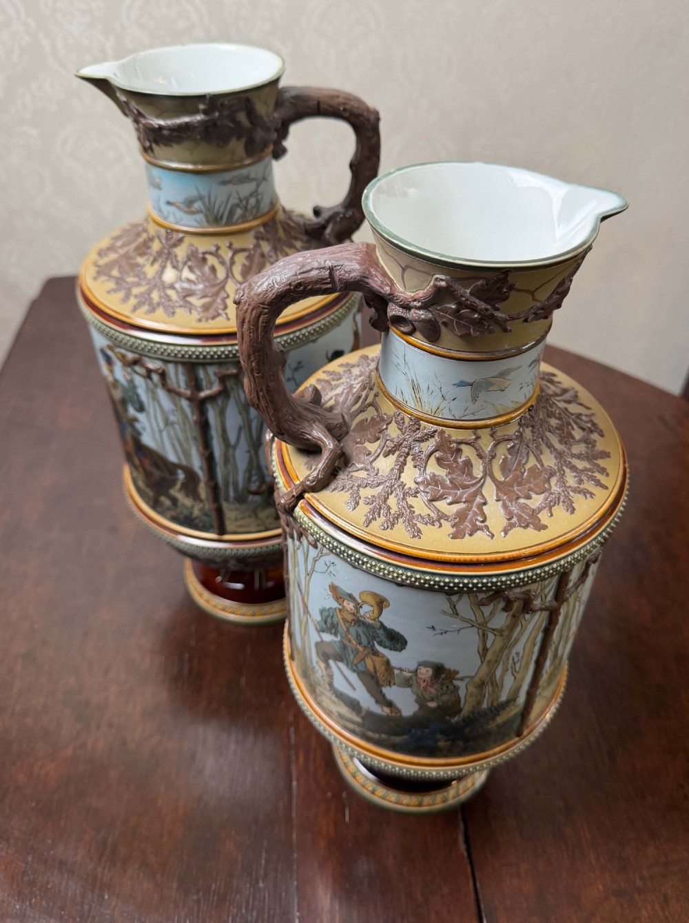 A pair of 19th-century Mettlach stoneware vases featuring sculpted branch handles, oak leaf relief decoration, and incised pictorial hunting scenes in blue and brown. View V