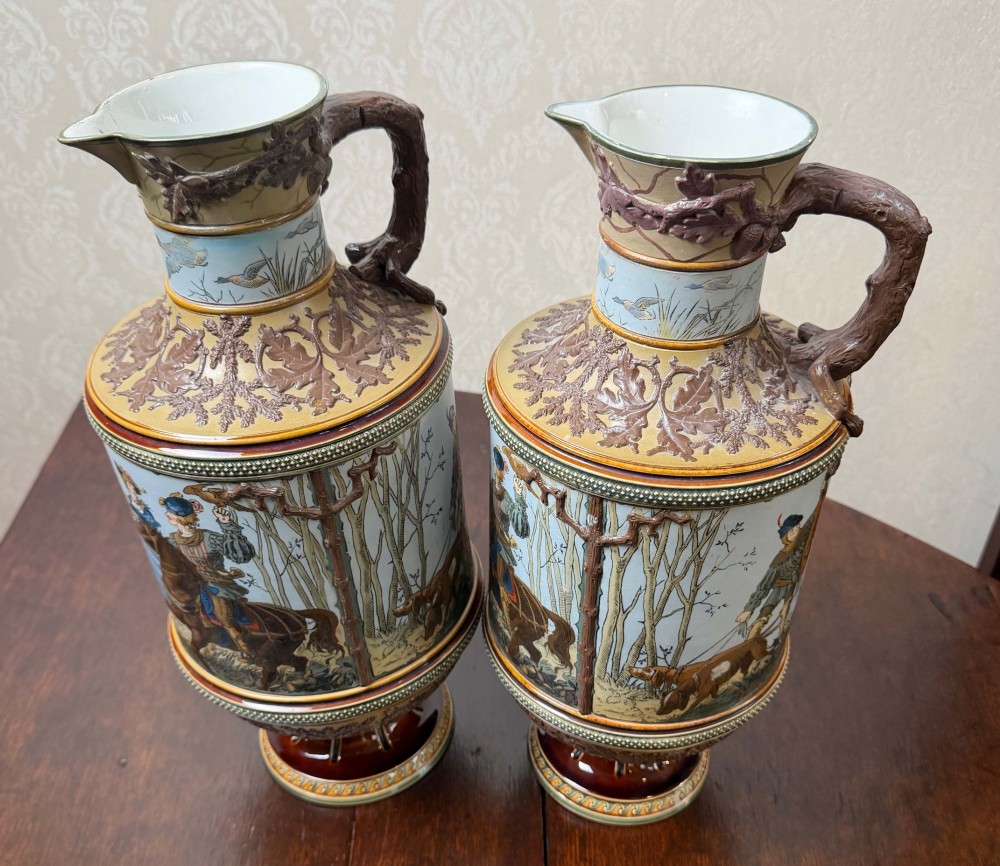 A pair of 19th-century Mettlach stoneware vases featuring sculpted branch handles, oak leaf relief decoration, and incised pictorial hunting scenes in blue and brown. View IX