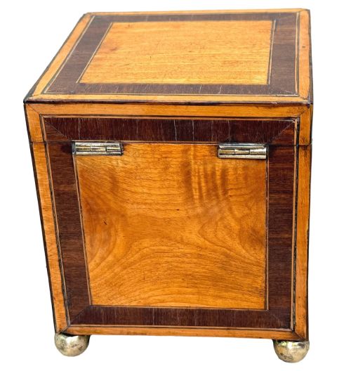 fig 2. A square Georgian satinwood tea caddy featuring dark wood crossbanded borders, a central brass keyhole escutcheon, and four brass ball feet.