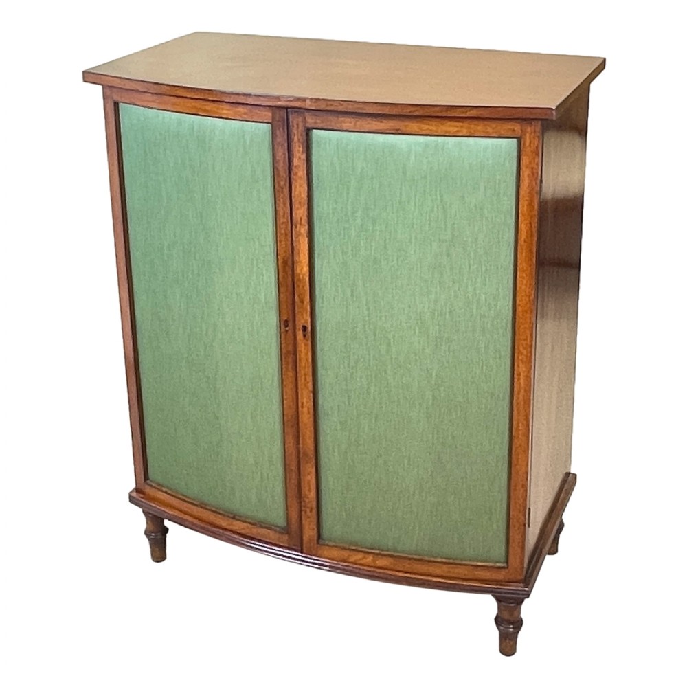 A Regency period bow-fronted mahogany side cabinet featuring two green fabric-panelled doors and turned legs.