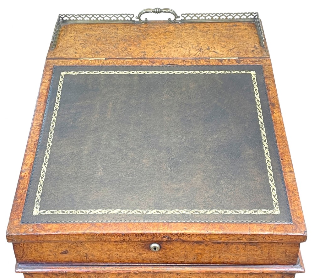 A Regency period burr elm Davenport desk featuring a sloped leather writing surface, a brass gallery railing, side drawers, bead moulding, and turned bun feet. View V