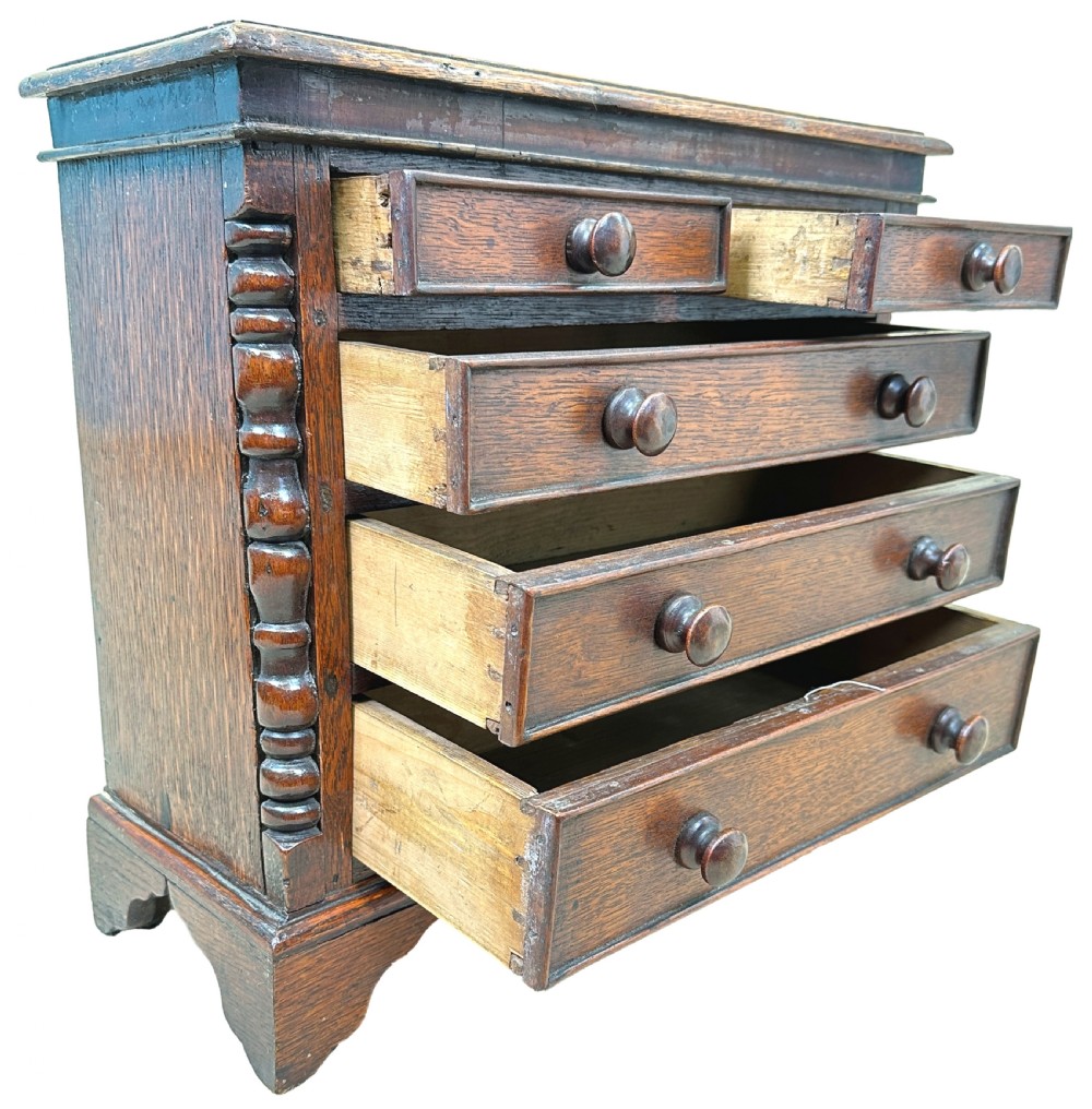 A Georgian oak miniature chest of drawers featuring two short over three long drawers with wooden knobs, turned half-columns, and bracket feet. View VI
