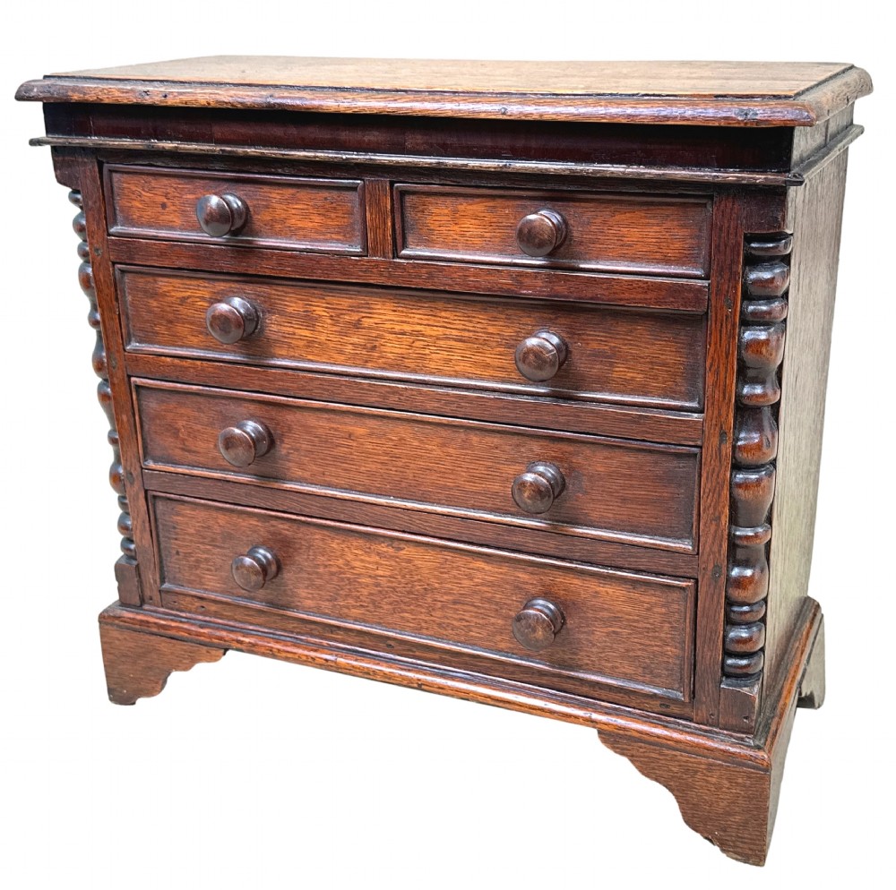 A Georgian oak miniature chest of drawers featuring two short over three long drawers with wooden knobs, turned half-columns, and bracket feet.