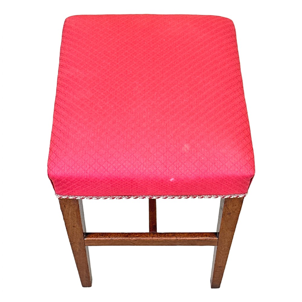 An 18th-century Georgian mahogany stool featuring a square red patterned upholstered top with braided trim, supported by four tapered wood legs and an H-stretcher. View IV