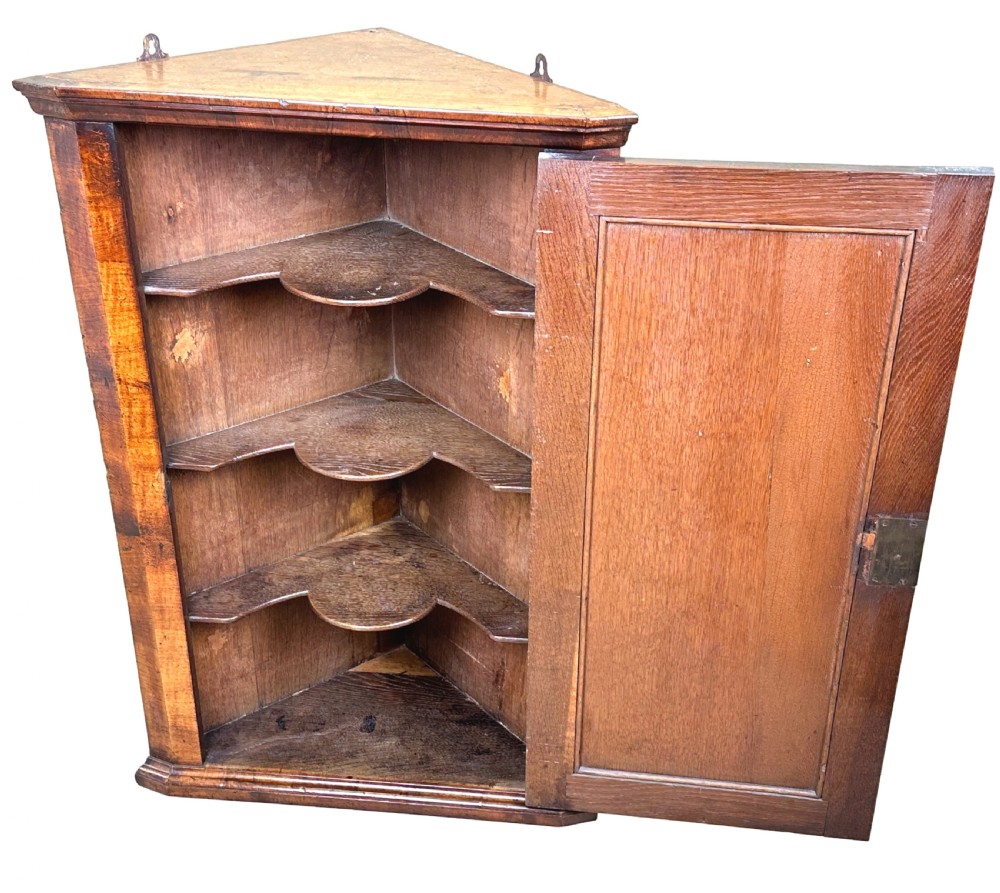 An early 18th-century walnut corner cupboard featuring a raised arched door panel, engraved brass H-hinges, and a moulded cornice. View IV