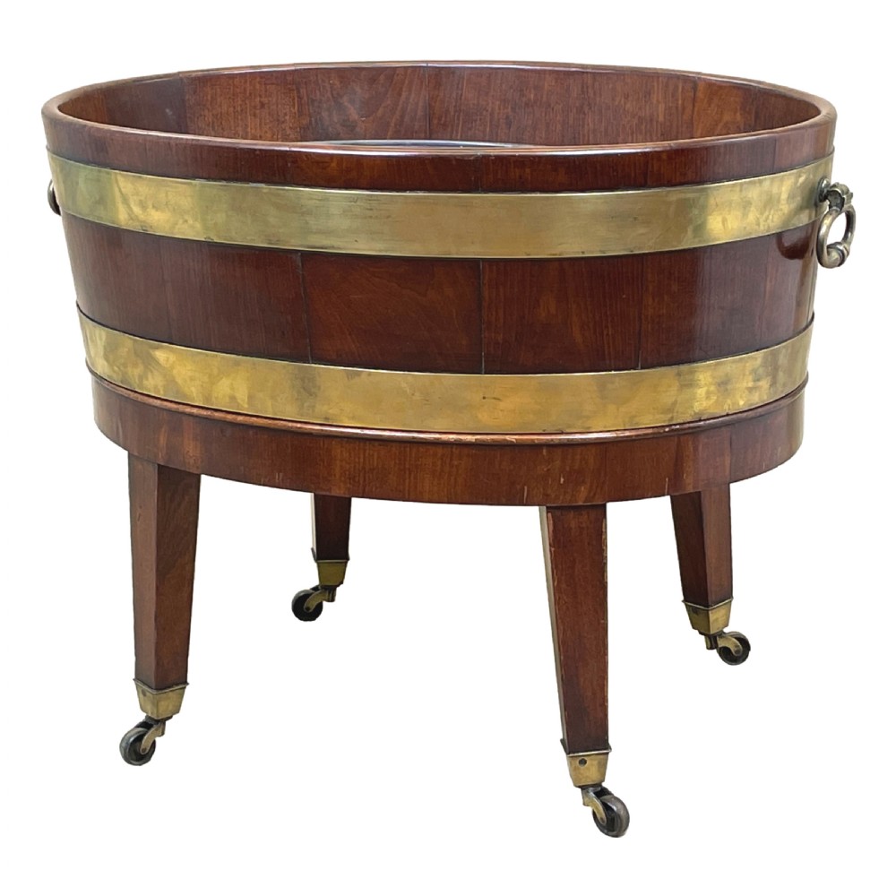 An oval coopered mahogany wine cooler featuring two brass binding hoops, carrying handles, and four tapered legs with brass castors.