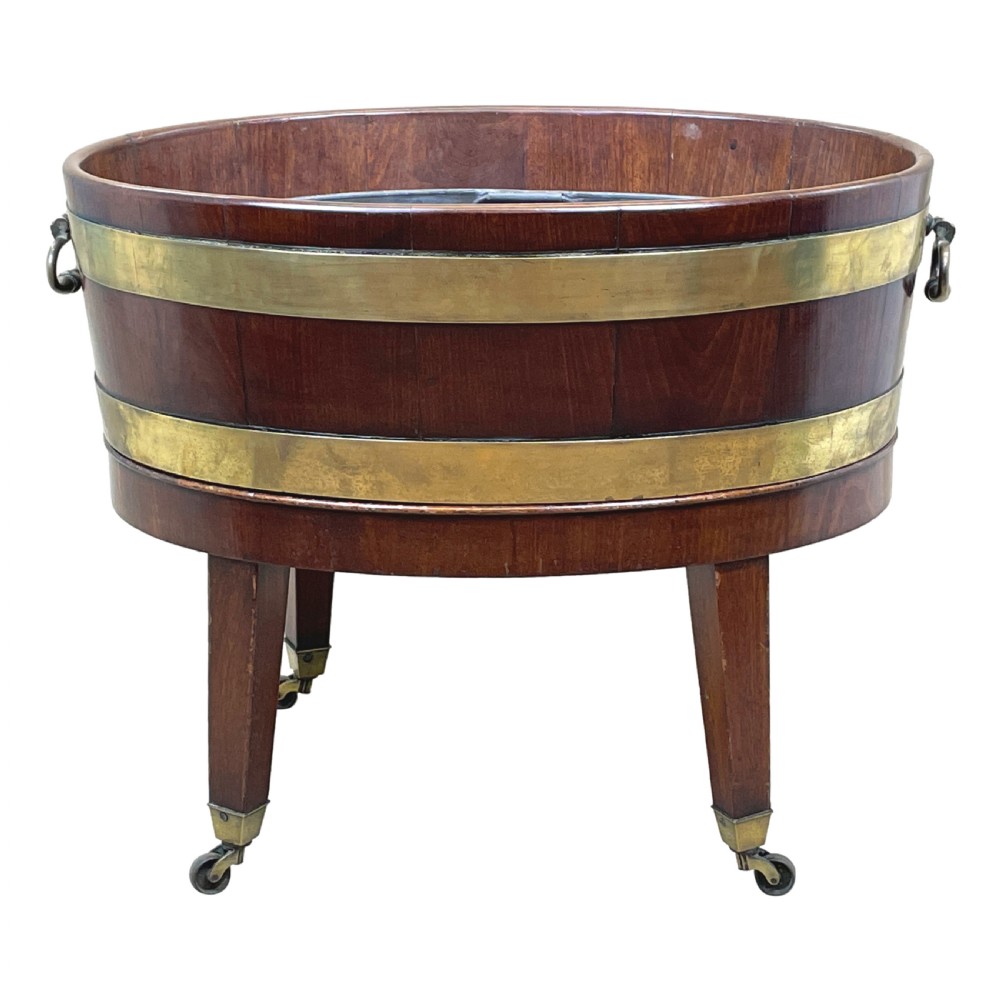 An oval coopered mahogany wine cooler featuring two brass binding hoops, carrying handles, and four tapered legs with brass castors. View V
