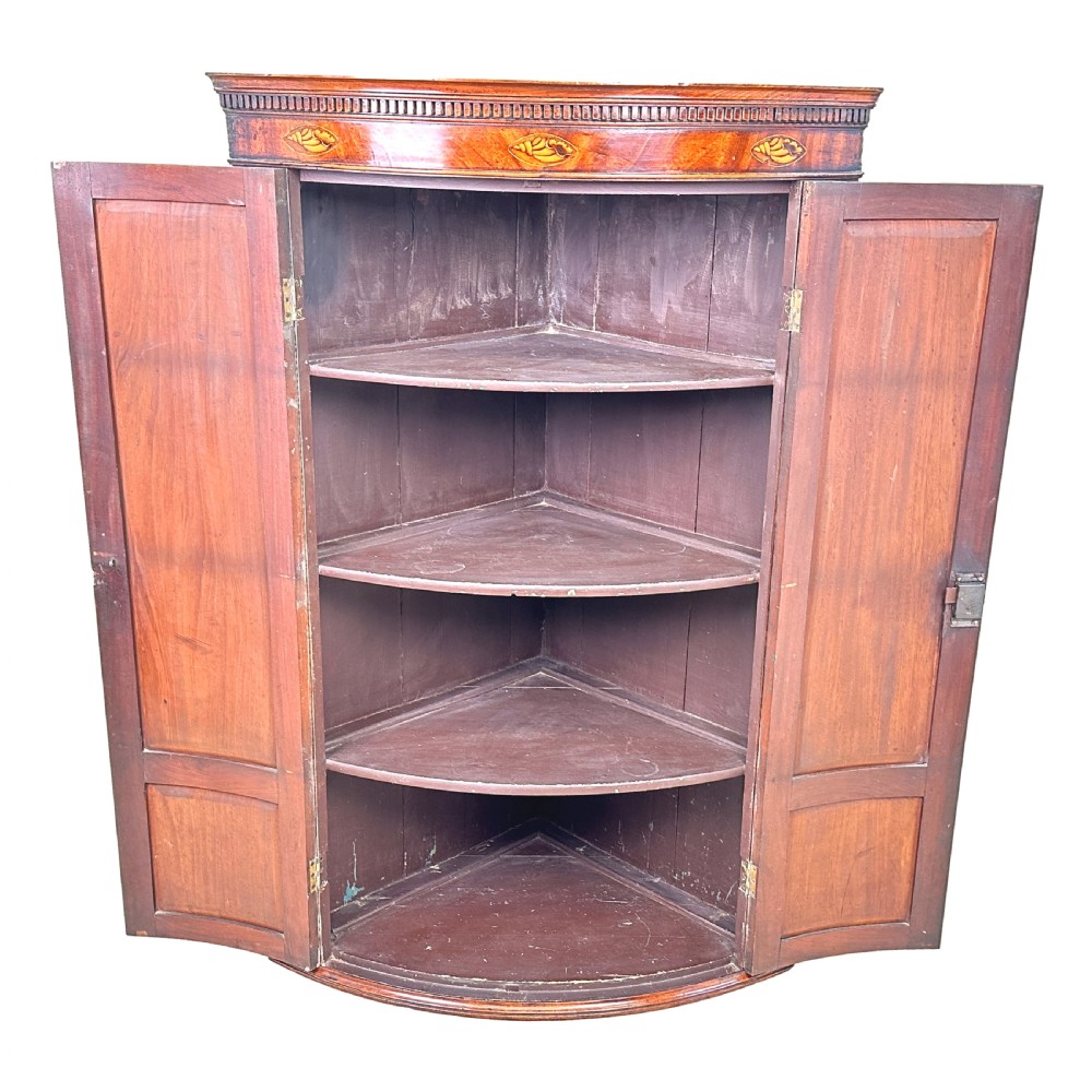 An 18th-century bow-front mahogany corner cupboard featuring double panelled doors, flame veneer, brass keyhole escutcheons, and inlaid shell motifs along the top frieze. View VII
