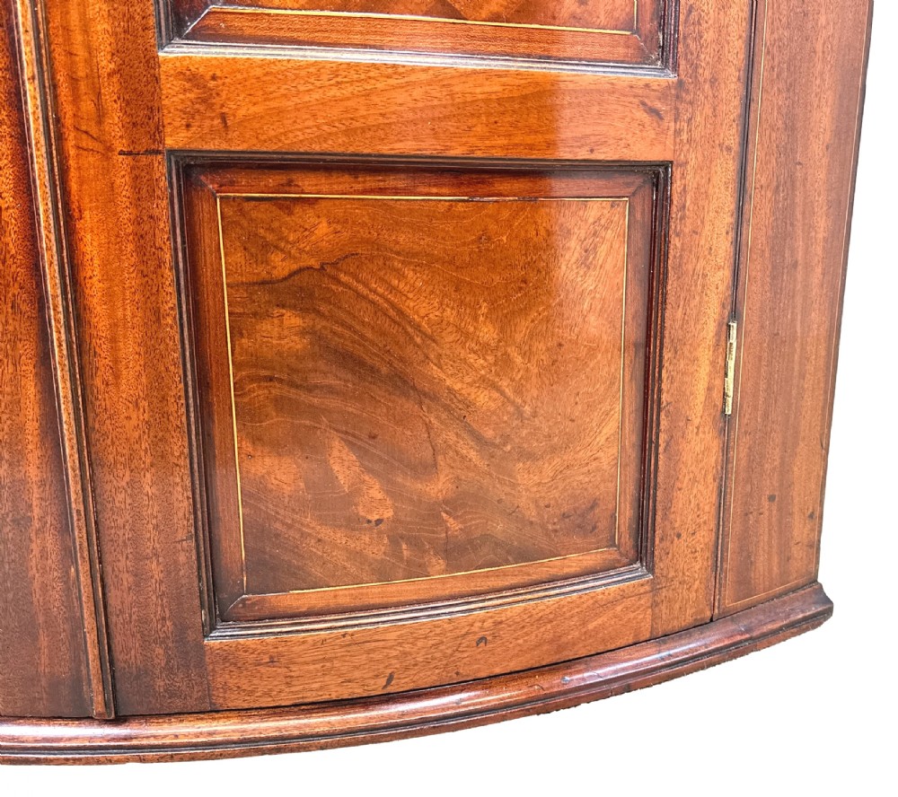 An 18th-century bow-front mahogany corner cupboard featuring double panelled doors, flame veneer, brass keyhole escutcheons, and inlaid shell motifs along the top frieze. View VIII