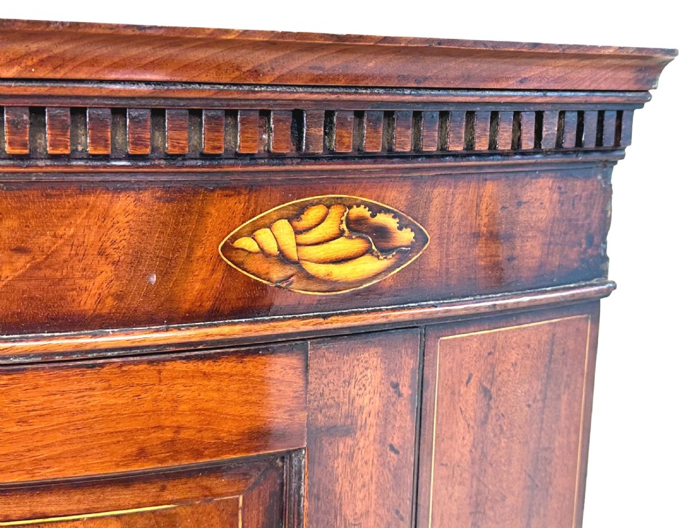 An 18th-century bow-front mahogany corner cupboard featuring double panelled doors, flame veneer, brass keyhole escutcheons, and inlaid shell motifs along the top frieze. View X