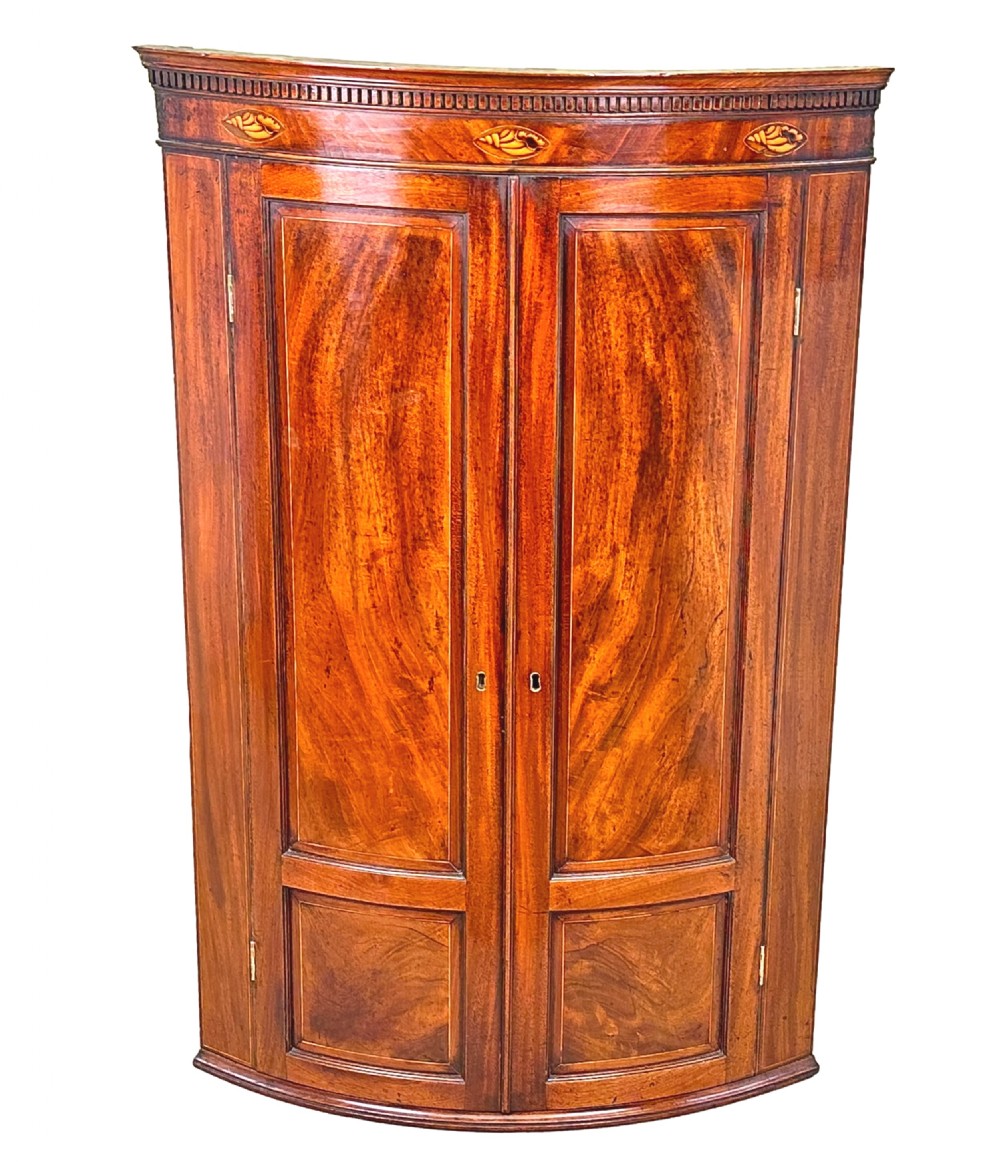 An 18th-century bow-front mahogany corner cupboard featuring double panelled doors, flame veneer, brass keyhole escutcheons, and inlaid shell motifs along the top frieze. View XI