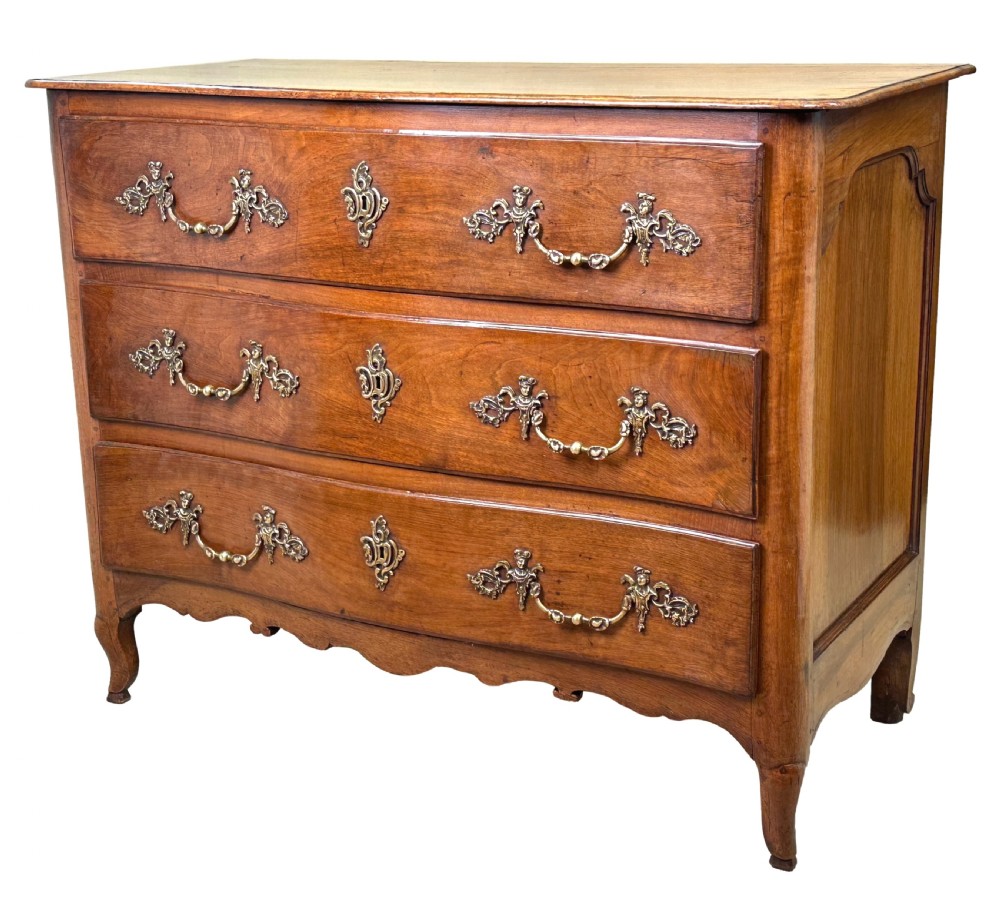 An 18th-century French provincial walnut commode chest featuring three full-width drawers decorated with elaborate brass handles and keyhole escutcheons, set above a scalloped apron and short cabriole legs.