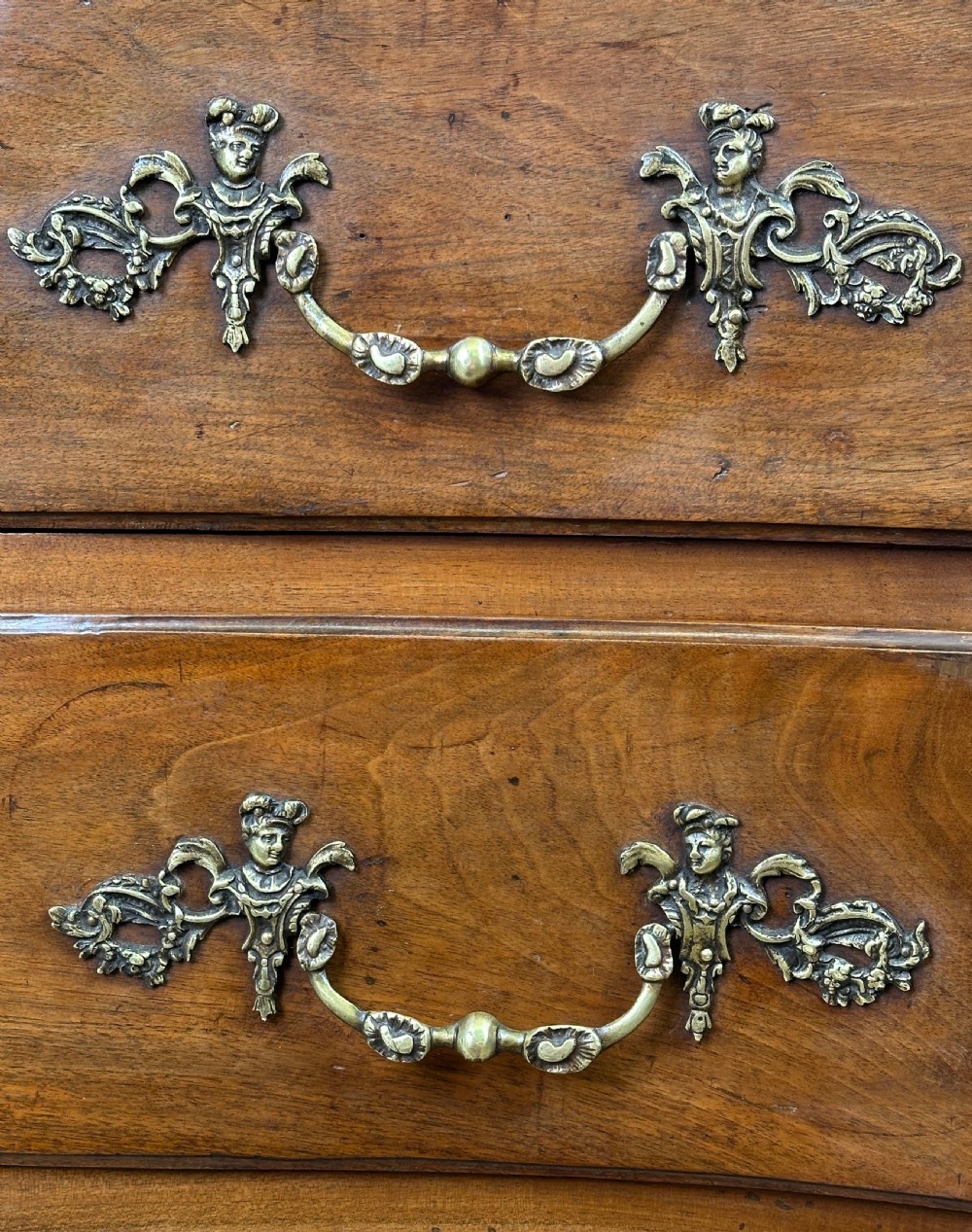 An 18th-century French provincial walnut commode chest featuring three full-width drawers decorated with elaborate brass handles and keyhole escutcheons, set above a scalloped apron and short cabriole legs. View X